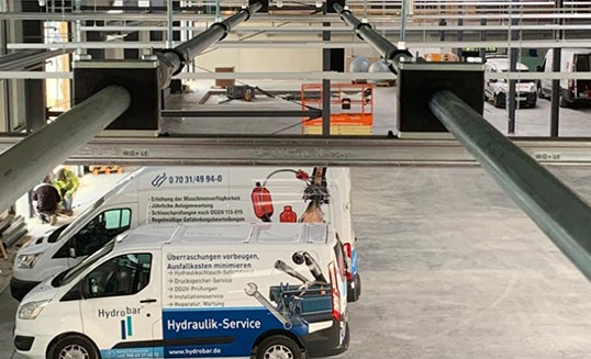 Hydraulische Hallenverrohrung mit großen Rohrleitungen über einer Industriehalle, darunter Servicefahrzeug von Hydrobar.